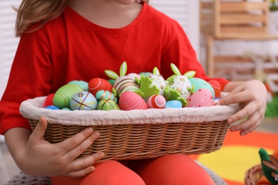 Photo of Little girl with wicker basket full of Easter eggs indoors, closeup