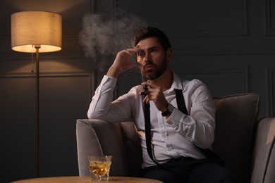 Photo of Tired man smoking cigar and resting at home in evening. Glass of whiskey on table near him
