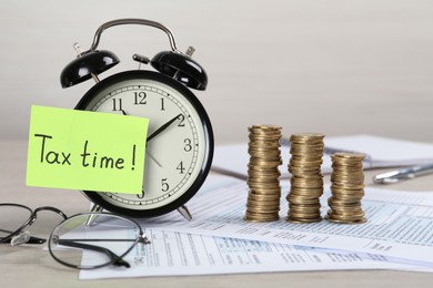 Photo of Alarm clock, reminder note with words Tax Time, documents and stacks of coins on white wooden table