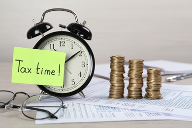 Alarm clock, reminder note with words Tax Time, documents and stacks of coins on white wooden table