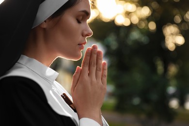 Young nun with hands clasped together praying outdoors on sunny day, closeup. Space for text