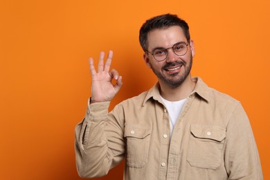 Photo of Portrait of smiling man in stylish glasses showing OK gesture on orange background. Space for text