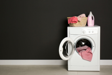 Modern washing machine with laundry, wicker basket and detergent near black wall. Space for text