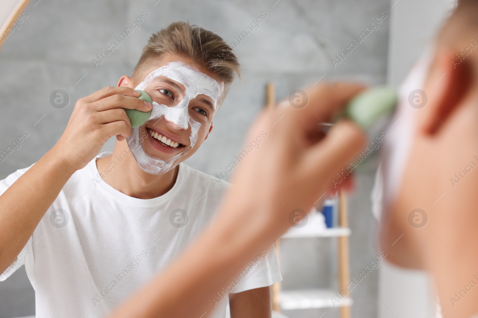 Photo of Happy young man washing off face mask with sponge near mirror in bathroom