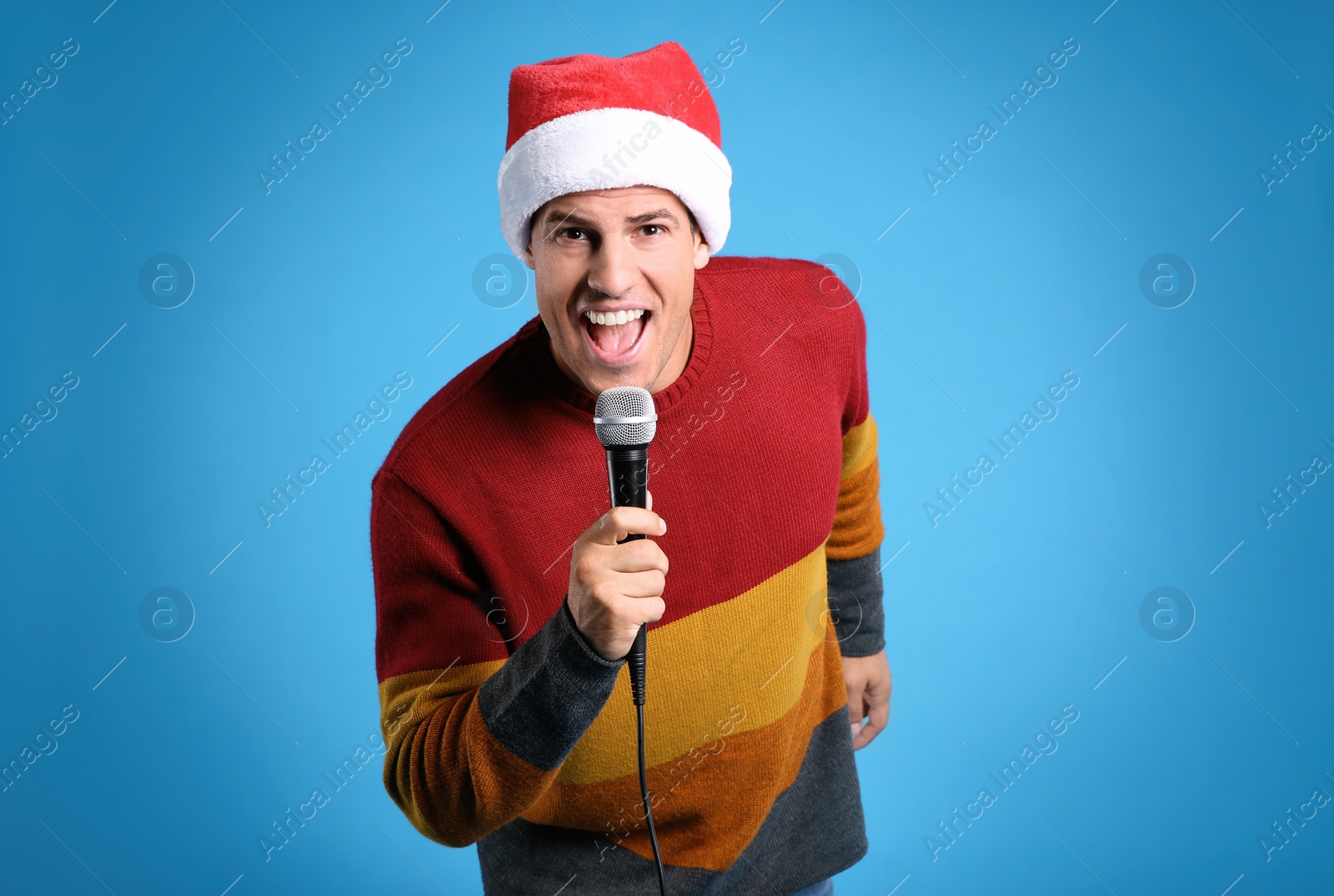 Photo of Emotional man in Santa Claus hat singing with microphone on blue background. Christmas music