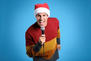 Emotional man in Santa Claus hat singing with microphone on blue background. Christmas music