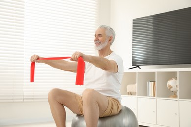 Photo of Senior man doing exercise with elastic resistance band on fitness ball at home