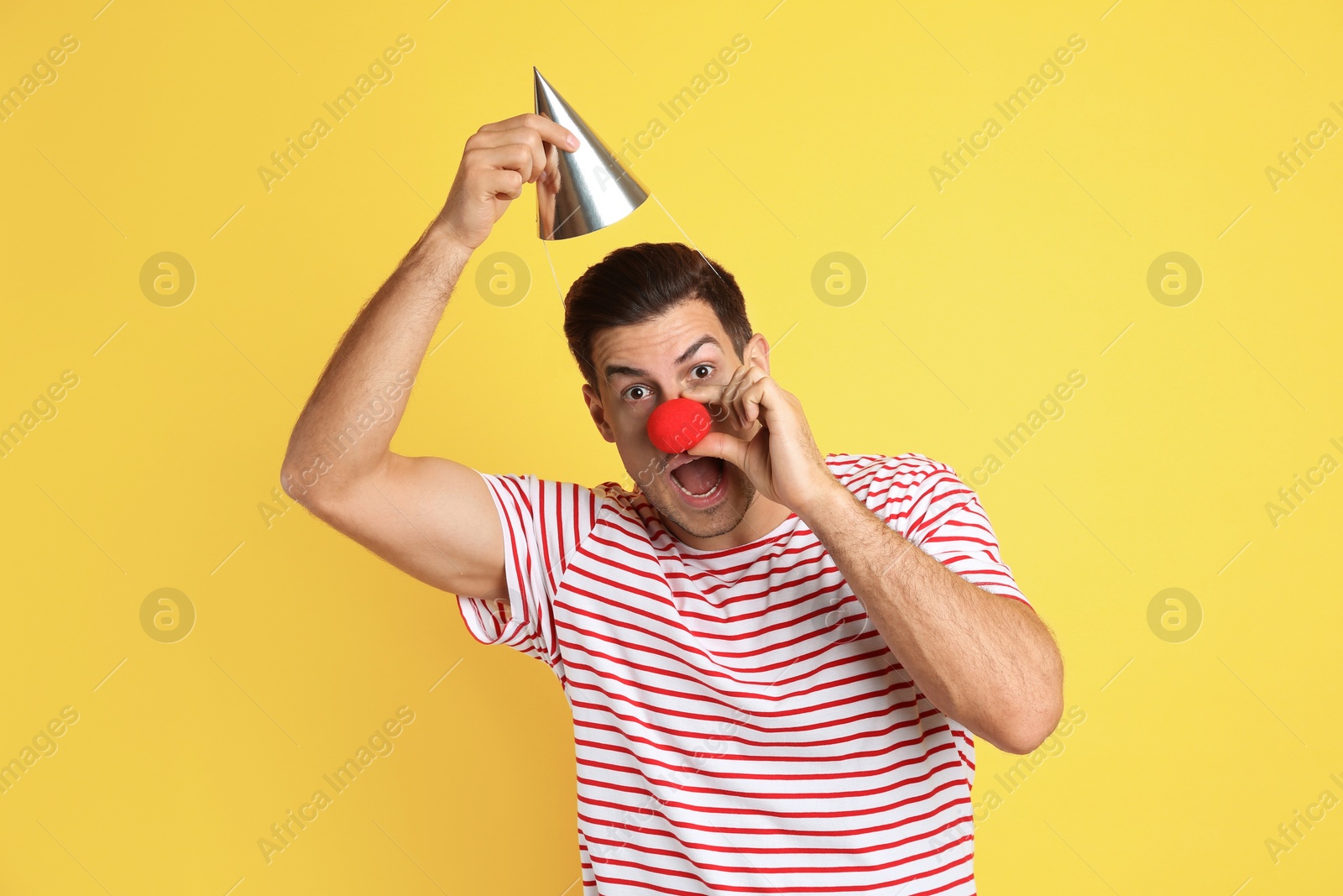 Photo of Funny man with clown nose and party hat on yellow background. April fool's day