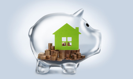 Transparent piggy bank with coins and house model on light background