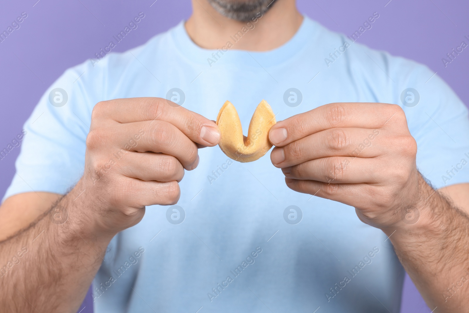 Photo of Man holding tasty fortune cookie with prediction on violet background, closeup