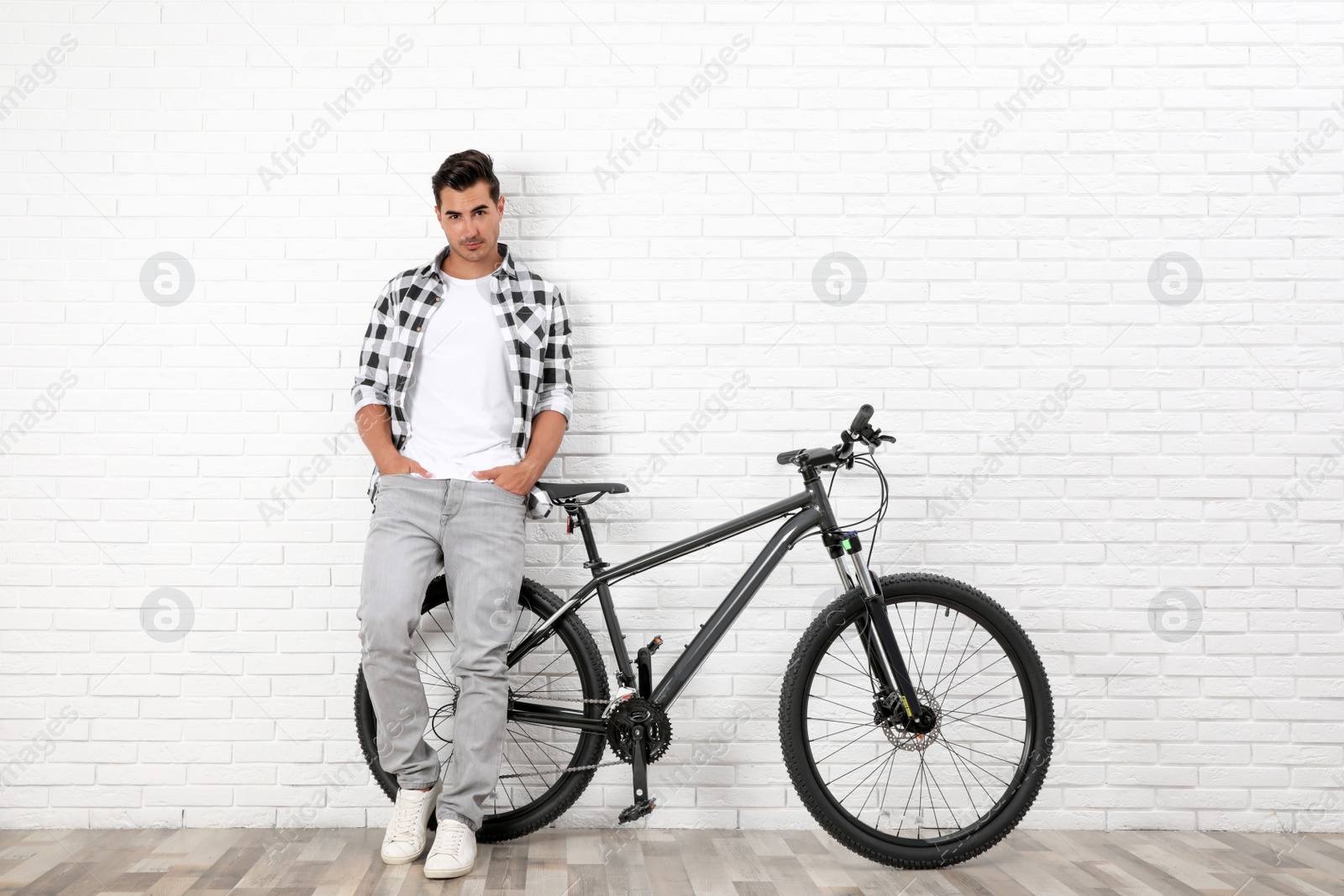 Photo of Handsome young man with modern bicycle near white brick wall indoors