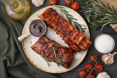 Photo of Tasty roasted pork ribs served with sauce, tomatoes and other products on grey wooden table, flat lay
