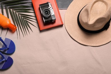 Beach towel, hat, book, camera, sunscreen, flip flops and tropical leaf on sand, flat lay. Space for text