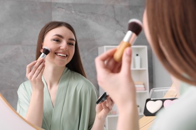 Photo of Beautiful young woman applying blusher with brush near mirror indoors