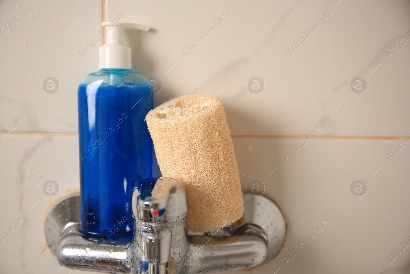 Photo of Natural loofah sponge and shower gel bottle on faucet in bathroom, closeup