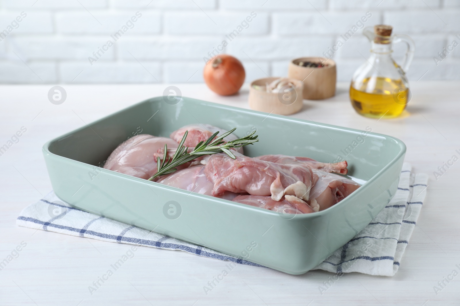 Photo of Raw rabbit meat and rosemary in baking dish on white wooden table