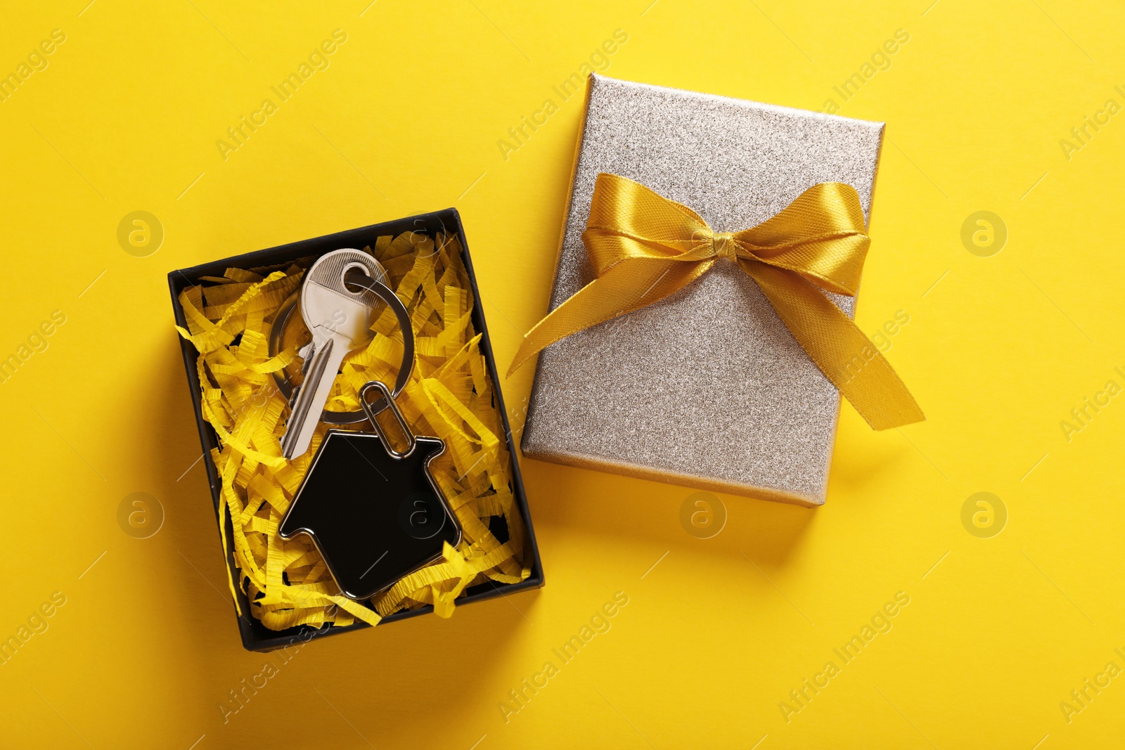 Photo of Key with trinket in shape of house and gift box on yellow background, flat lay. Housewarming party