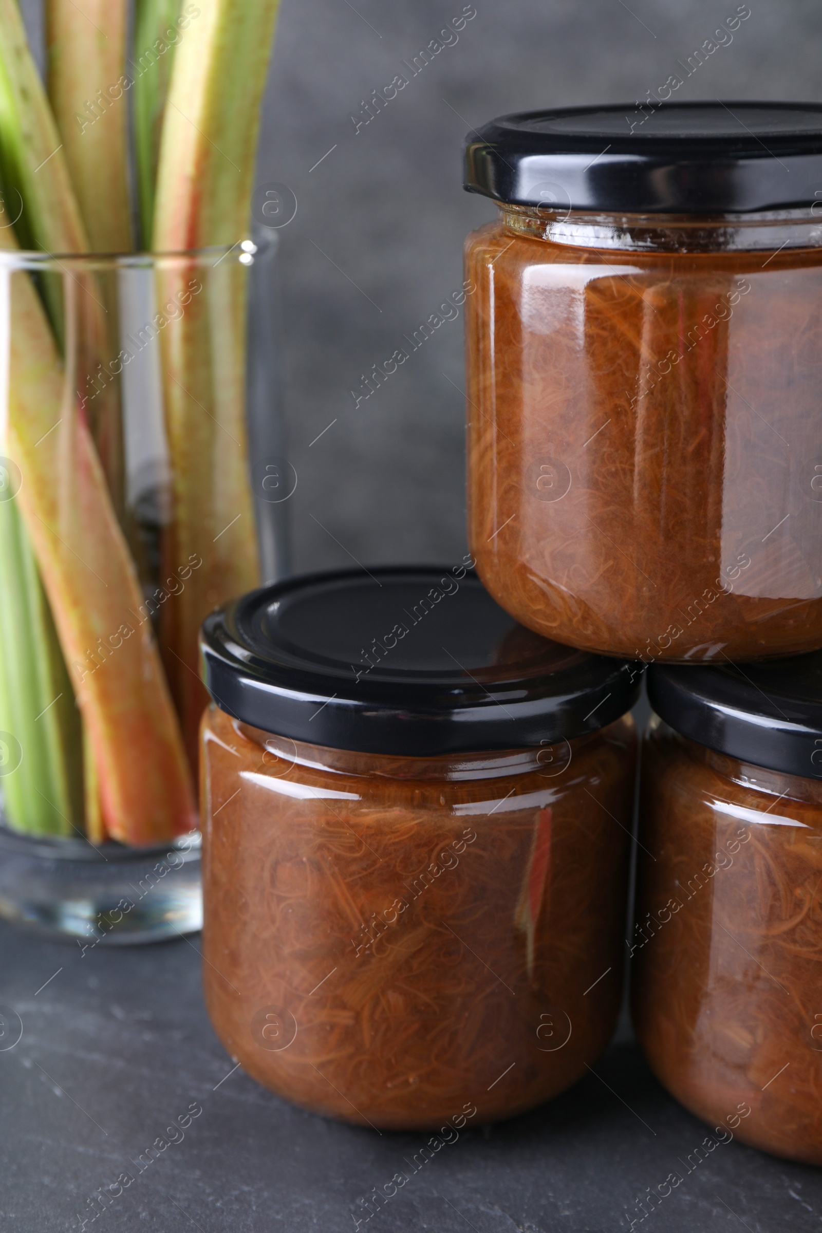 Photo of Jars of tasty rhubarb jam and stalks on grey table, closeup