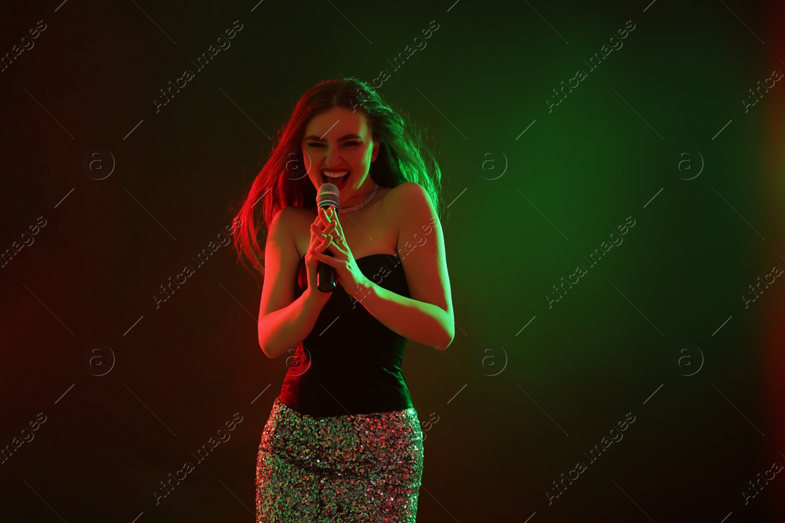 Photo of Emotional woman with microphone singing in color lights on dark background. Space for text