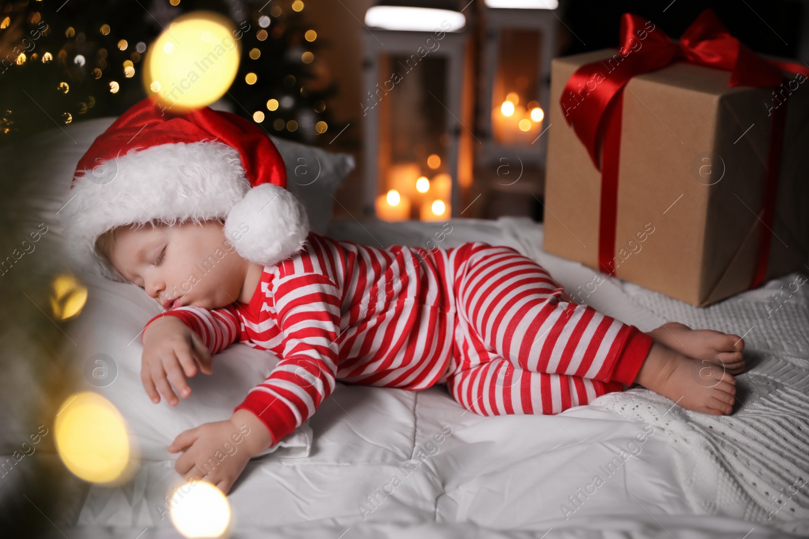 Photo of Baby in Christmas pajamas and Santa hat sleeping near gift box on bed indoors