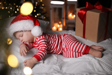 Photo of Baby in Christmas pajamas and Santa hat sleeping near gift box on bed indoors