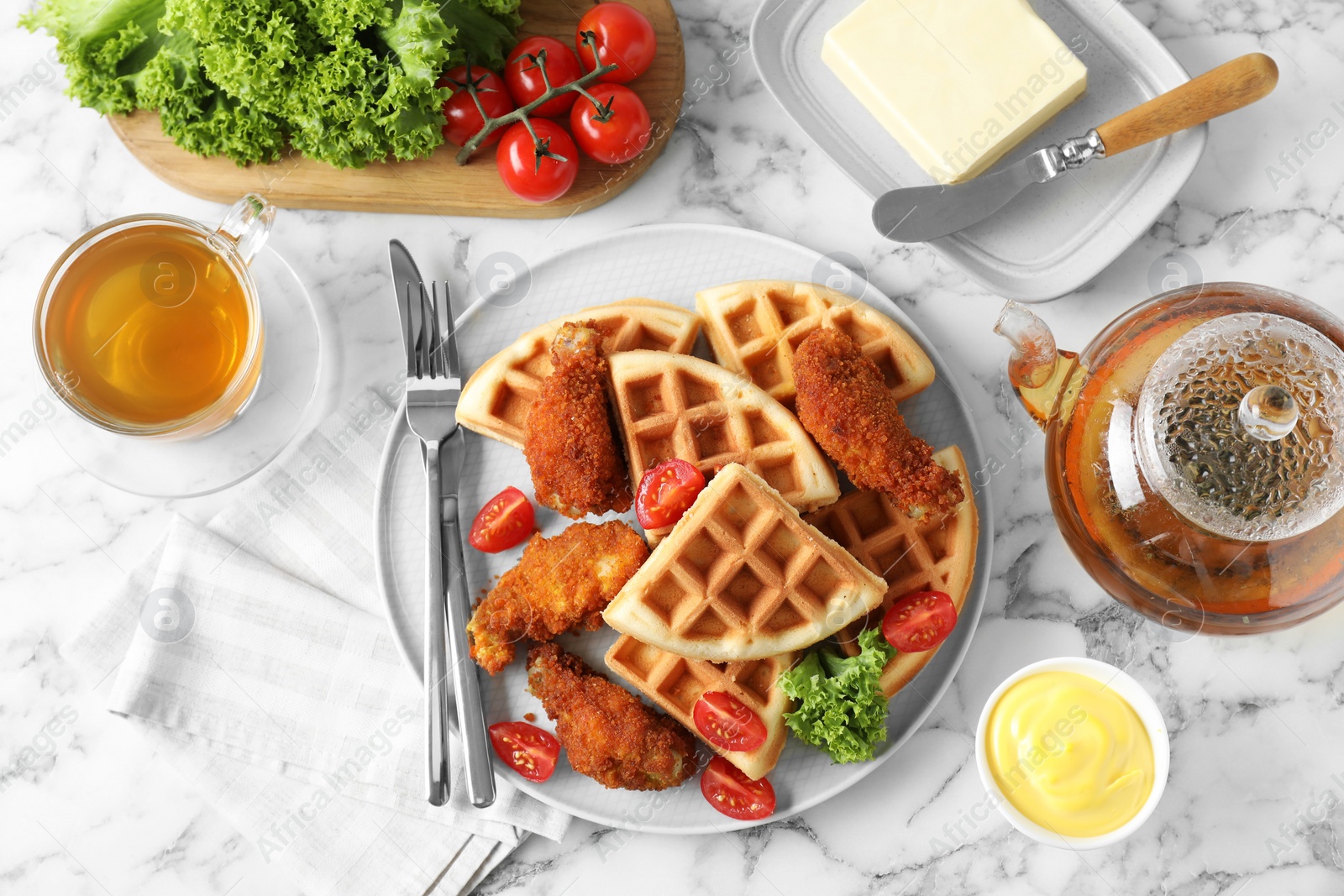 Photo of Tasty Belgian waffles served with fried chicken, tomatoes, lettuce and tea on white marble table, flat lay