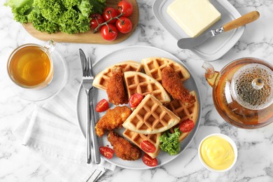 Tasty Belgian waffles served with fried chicken, tomatoes, lettuce and tea on white marble table, flat lay