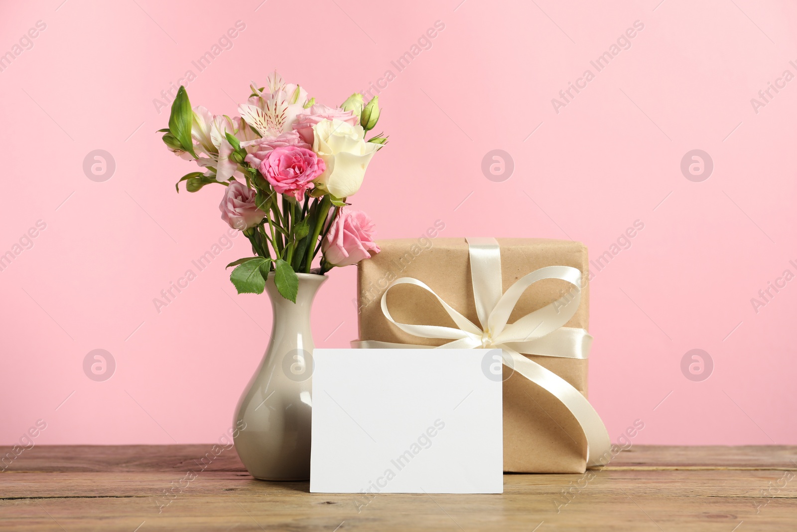 Photo of Happy Mother's Day. Gift box, blank card and bouquet of beautiful flowers in vase on wooden table against pink background