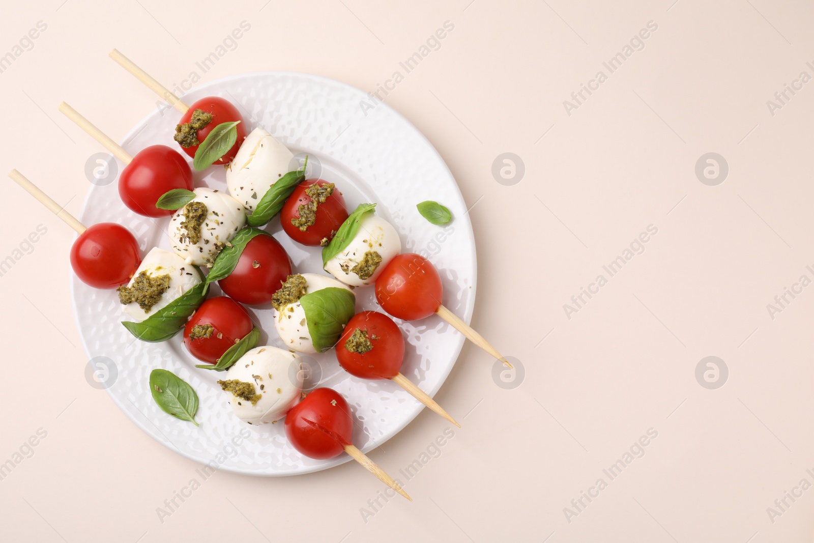 Photo of Caprese skewers with tomatoes, mozzarella balls, basil and pesto sauce on beige background, top view. Space for text