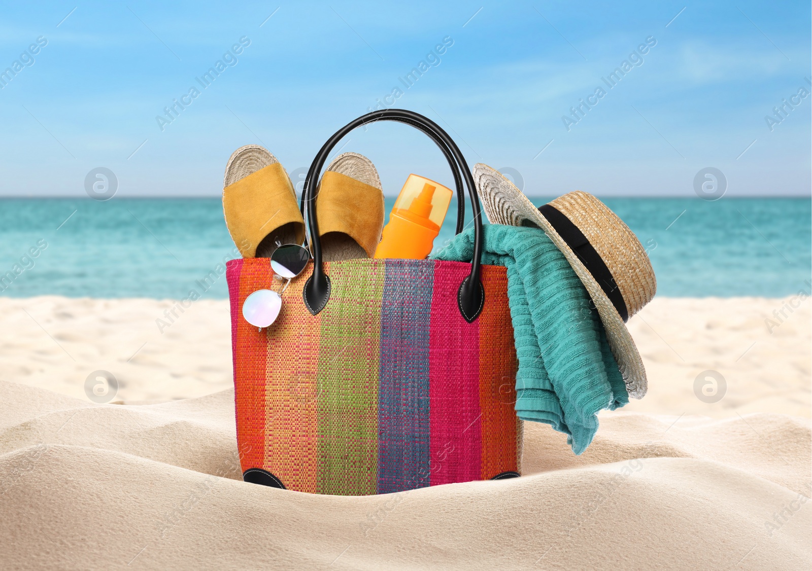 Image of Stylish bag with different accessories on sandy beach near ocean