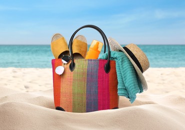 Stylish bag with different accessories on sandy beach near ocean