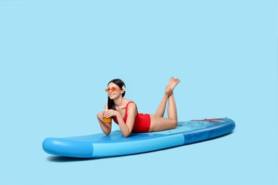 Happy woman with refreshing drink resting on SUP board against light blue background