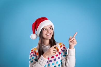 Photo of Pretty woman in Santa hat and festive sweater pointing on light blue background