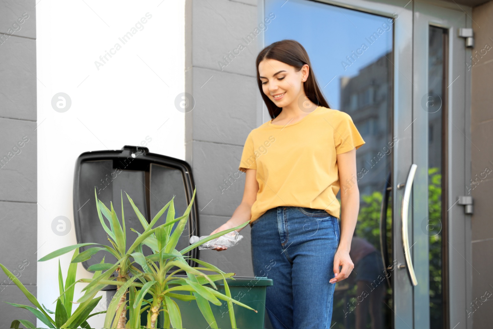 Photo of Young woman throwing plastic bottle into recycling bin outdoors