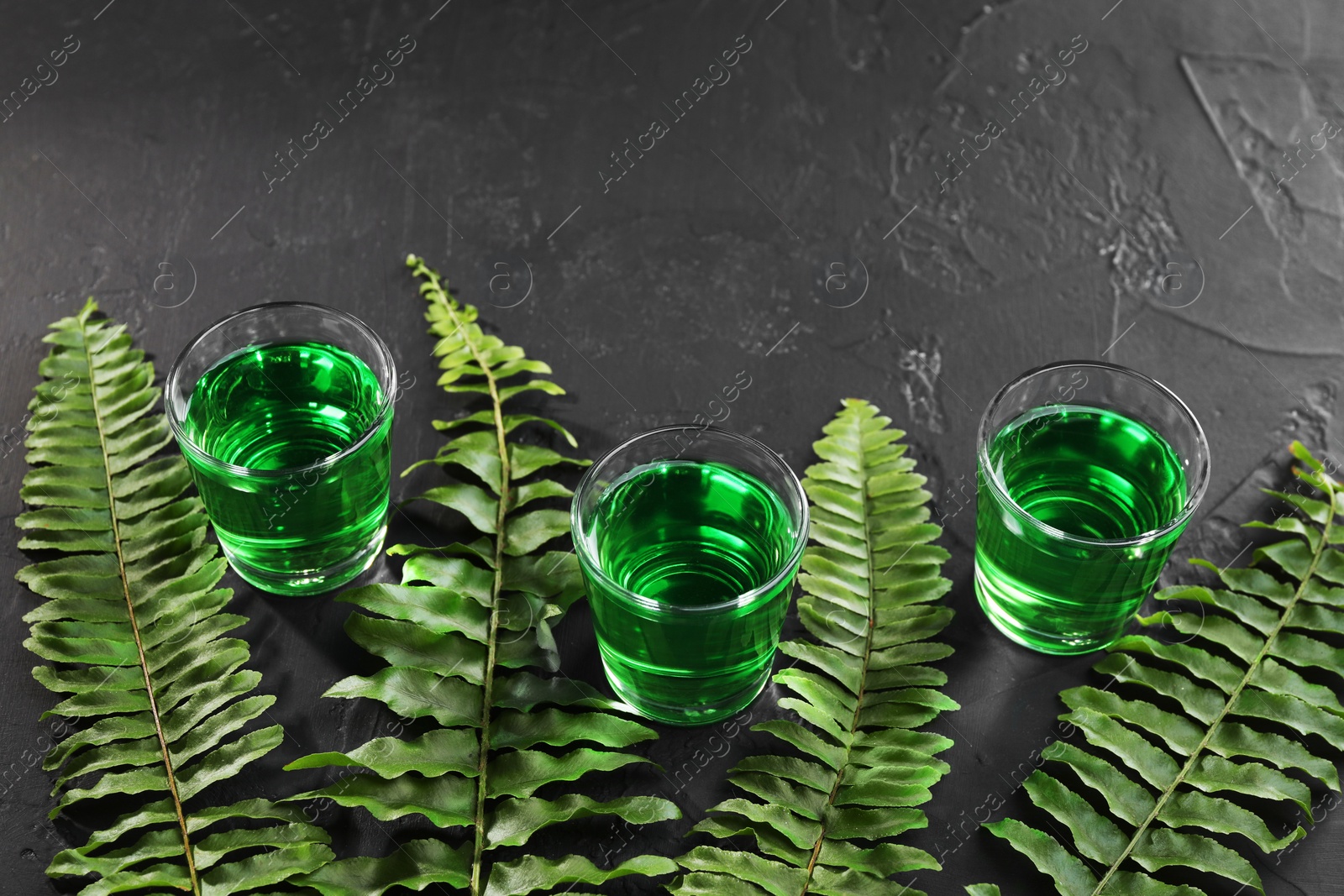 Photo of Absinthe in shot glasses and green leaves on black table, space for text. Alcoholic drink