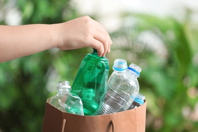 Photo of Woman putting used plastic bottle into paper bag on blurred background, closeup with space for text. Recycling problem