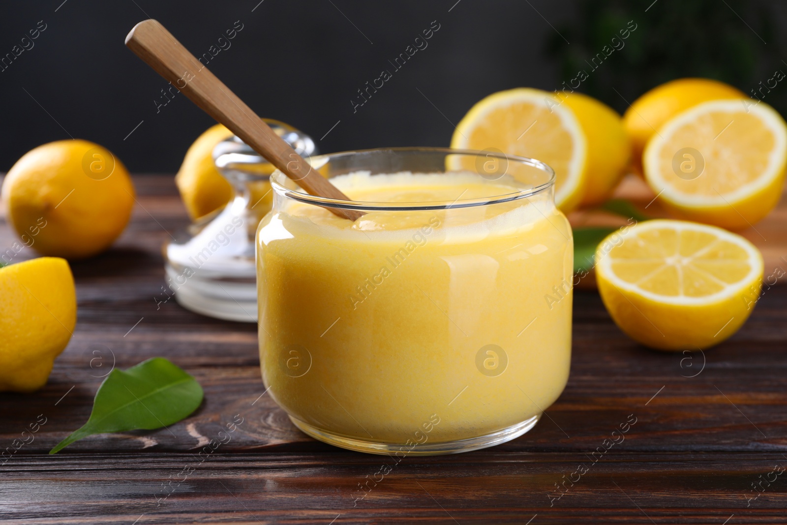 Photo of Delicious lemon curd in glass jar, fresh citrus fruits and spoon on wooden table