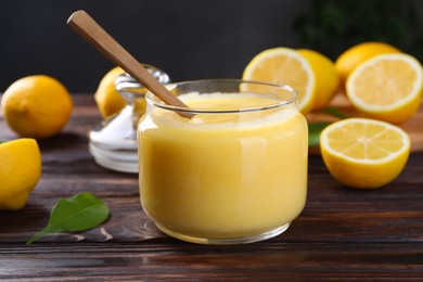 Photo of Delicious lemon curd in glass jar, fresh citrus fruits and spoon on wooden table