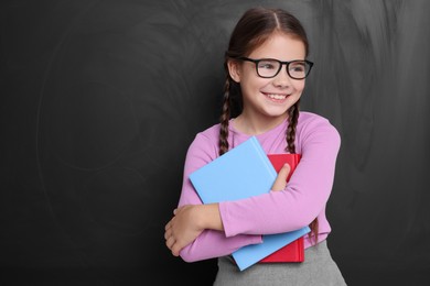 Cute schoolgirl in glasses holding books near chalkboard, space for text