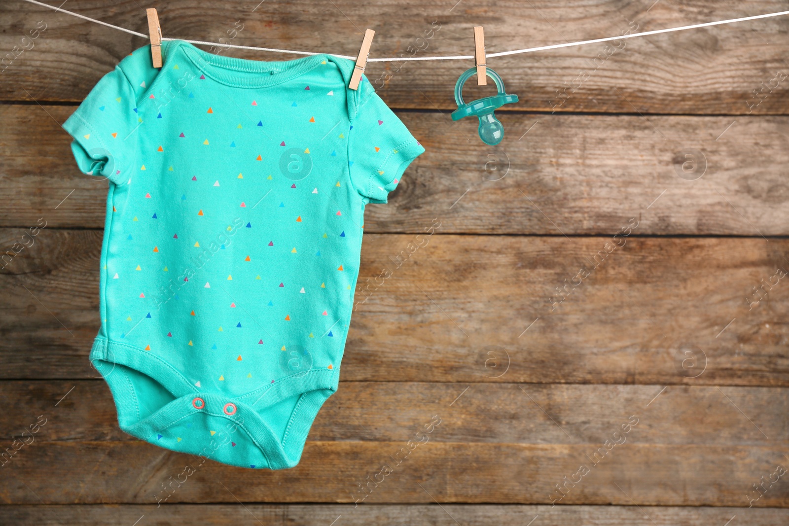 Photo of Bodysuit and pacifier on laundry line against wooden background, space for text. Baby accessories