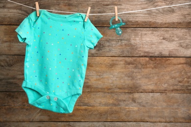 Photo of Bodysuit and pacifier on laundry line against wooden background, space for text. Baby accessories