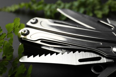 Photo of Compact portable metallic multitool and green leaves on black background, closeup