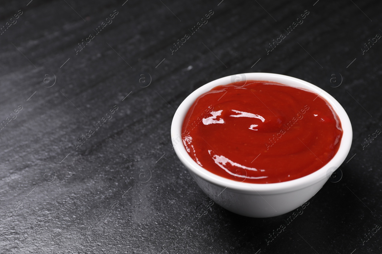 Photo of Organic ketchup in bowl on black table, space for text. Tomato sauce