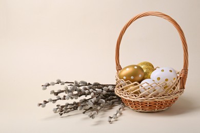 Photo of Wicker basket with decorated Easter eggs and willow branches on beige background. Space for text