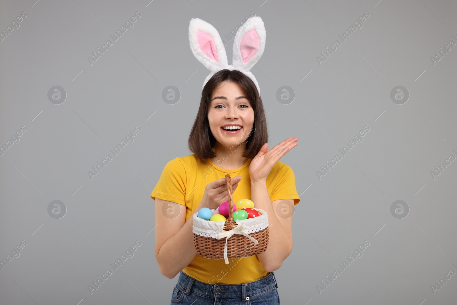 Photo of Easter celebration. Happy woman with bunny ears and wicker basket full of painted eggs on grey background