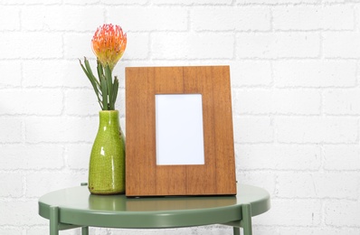 Photo of Blank frame and vase with flower on table near brick wall. Mock up for design