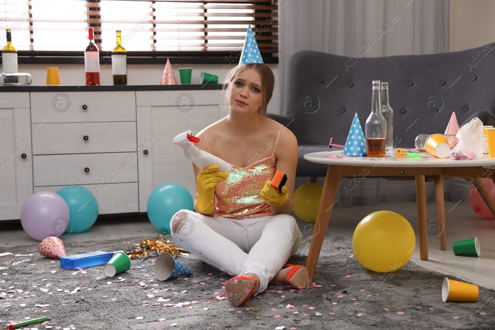 Photo of Tired young woman with bottle of detergent and sponge in messy room after party