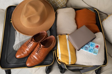 Open suitcase with packed clothes, accessories and pill box on sofa, above view