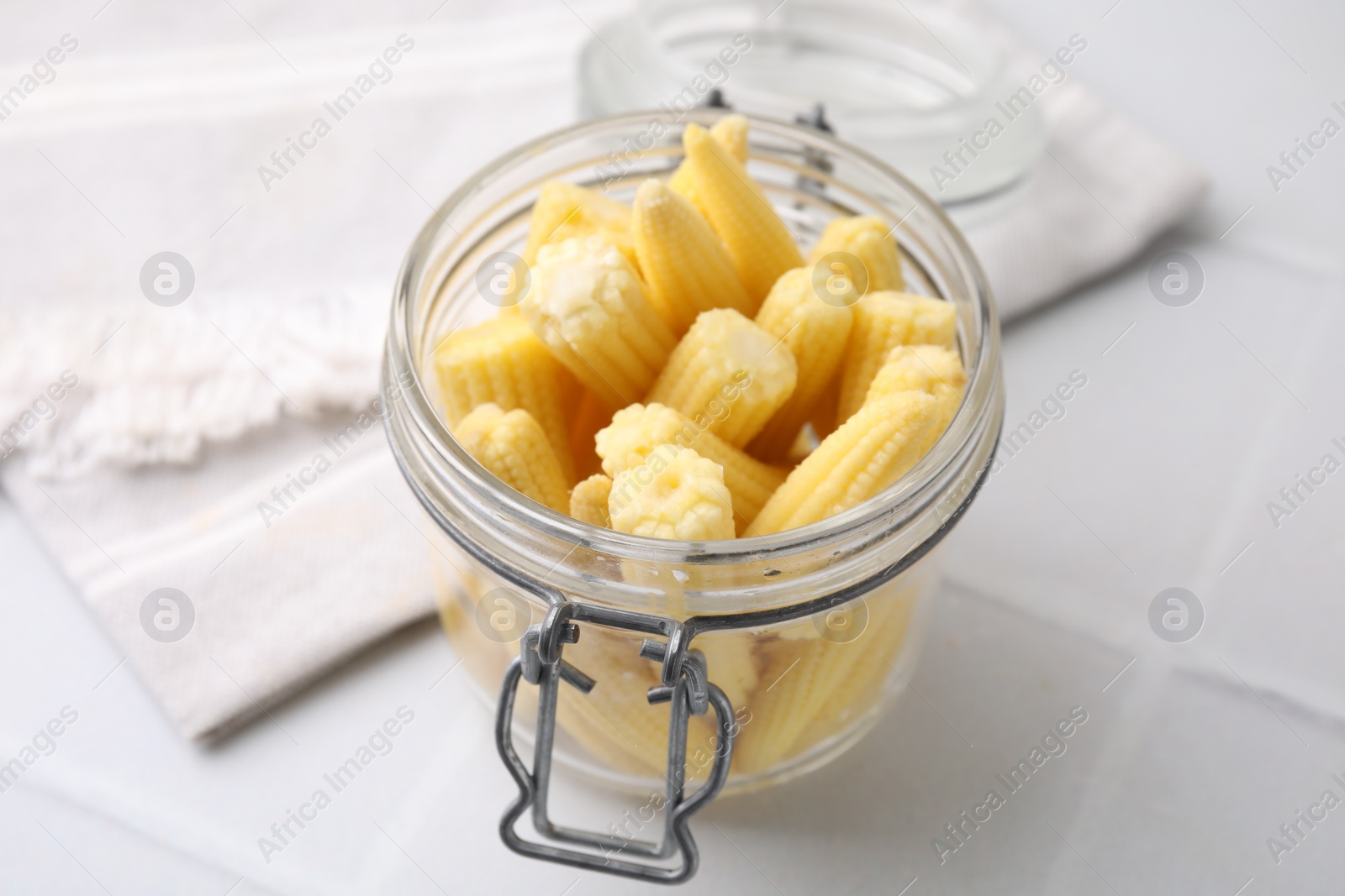 Photo of Tasty fresh yellow baby corns in glass jar on white tiled table, closeup