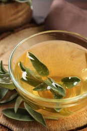 Photo of Cup of aromatic sage tea and fresh leaves on wooden stump, closeup